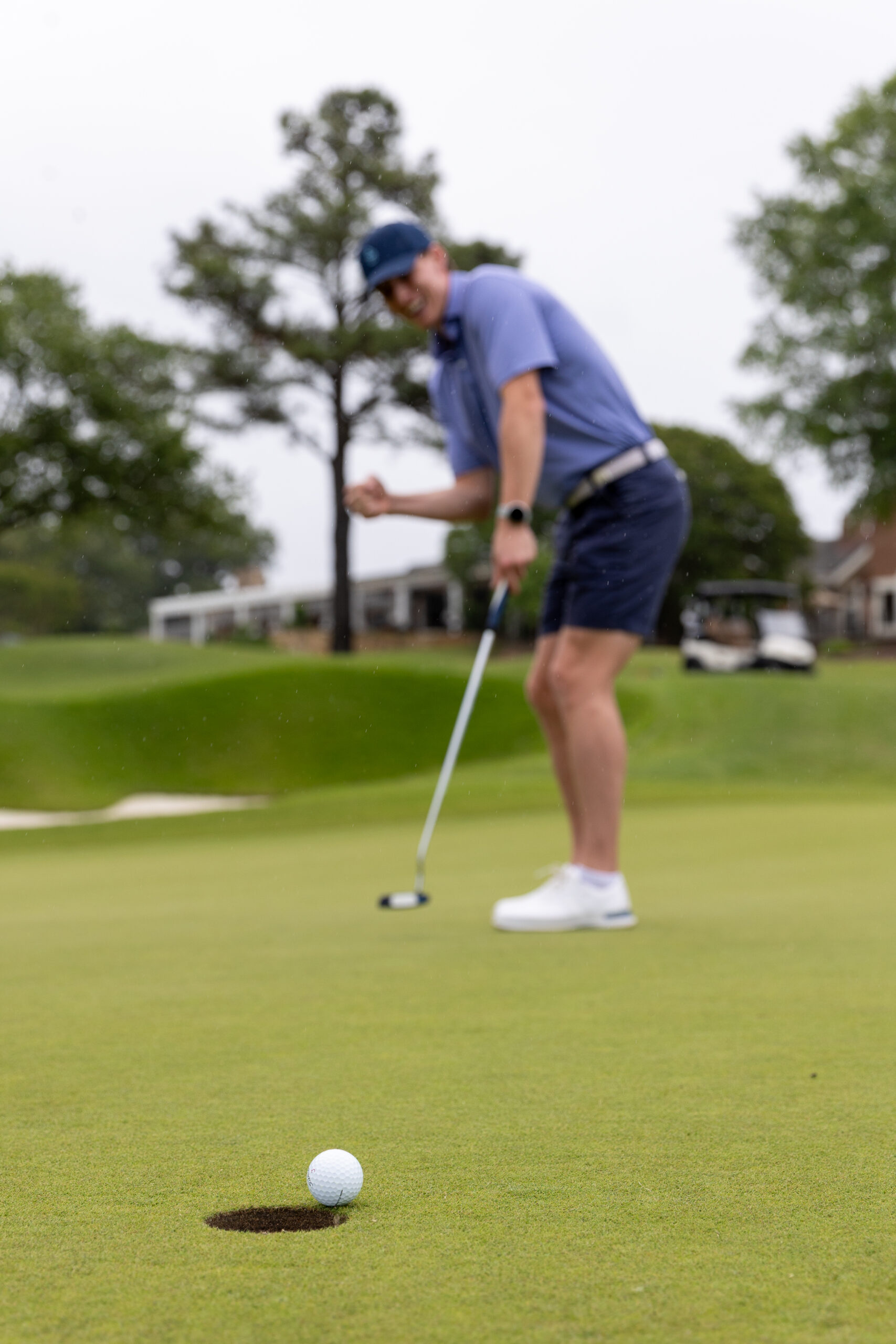 A golfer in a blue shirt and shorts celebrates with a fist pump as his golf ball stops just short of the hole on a green—capturing the excitement found at events for Virginia boys and girls, with trees and a golf cart in the background.