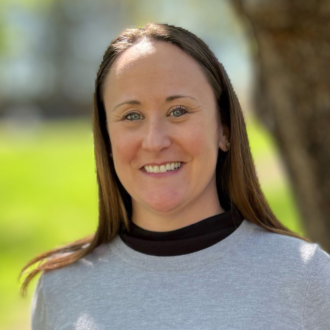 A woman with straight brown hair smiles outdoors, wearing a light gray sweater over a black top. The blurred background suggests green grass, a tree trunk, and sunlight—reflecting the welcoming environment of Virginia education group homes.