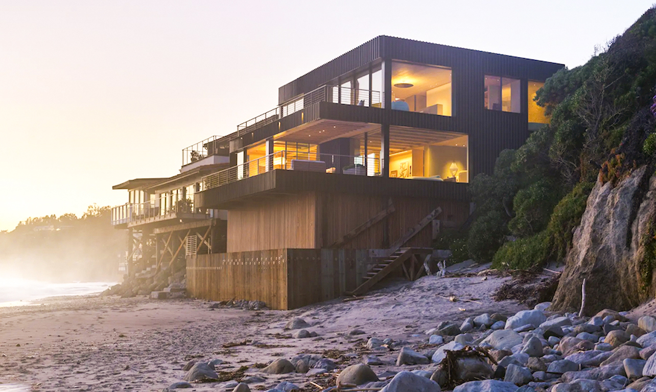 Modern beachfront house with large windows and balconies, elevated on wooden supports, overlooking a rocky, sandy beach at sunset. Warm interior lights contrast with the natural outdoor setting.
