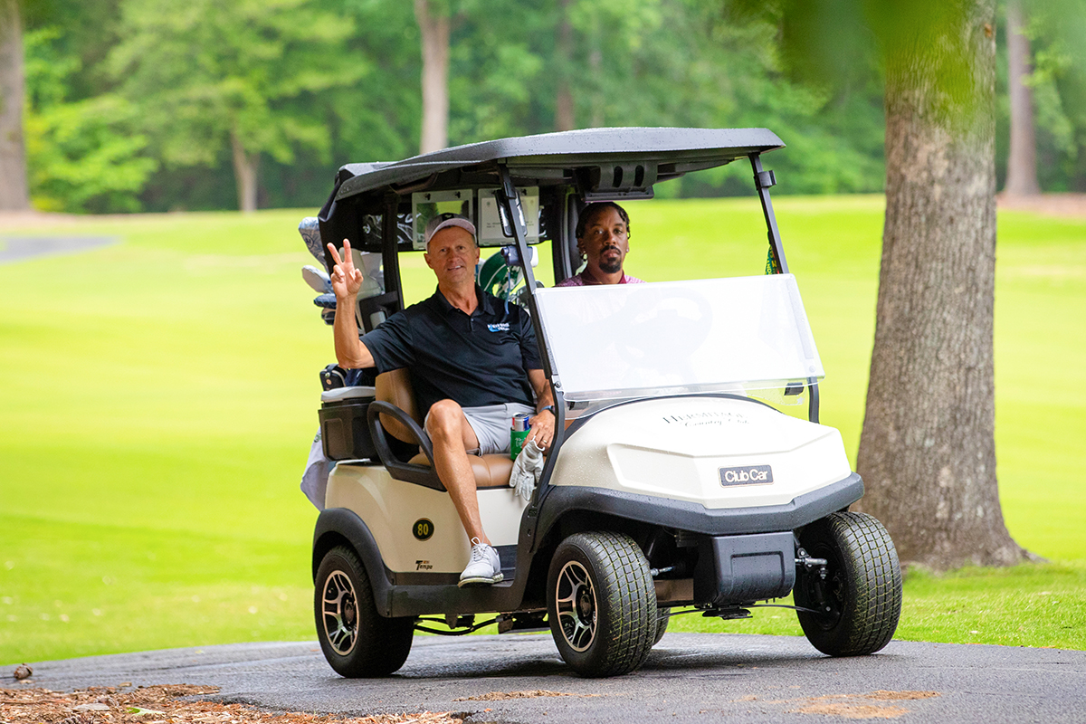 Two men ride in a golf cart on a green golf course, enjoying events for Virginia boys and girls. One man, wearing a cap and shorts, smiles and makes a peace sign, while the other sits beside him. Trees and grass fill the background.