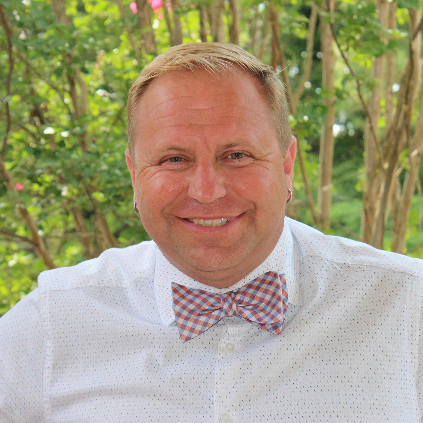 A smiling man with short blond hair wearing a white shirt with small blue dots and a pink and blue plaid bow tie stands outdoors in front of green trees, representing Virginia education group homes services for boys and girls.