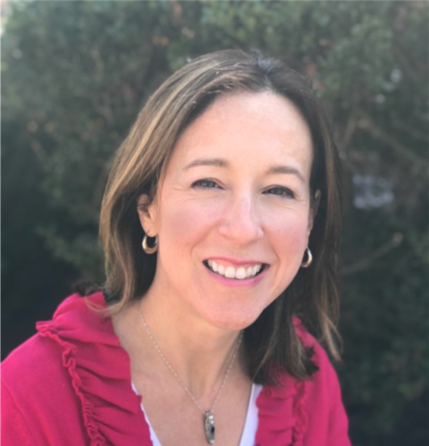 A woman with shoulder-length brown hair smiles at the camera. She is wearing a pink top with ruffles, hoop earrings, and a pendant necklace. Blurred greenery is visible in the background at a Virginia education group home for boys and girls.