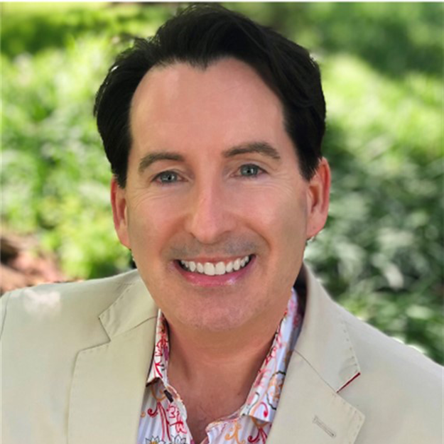 A smiling man with dark hair wears a beige blazer and a colorful patterned shirt, posing outdoors with green foliage in the background. He works with Virginia education group homes, offering services for boys and girls.