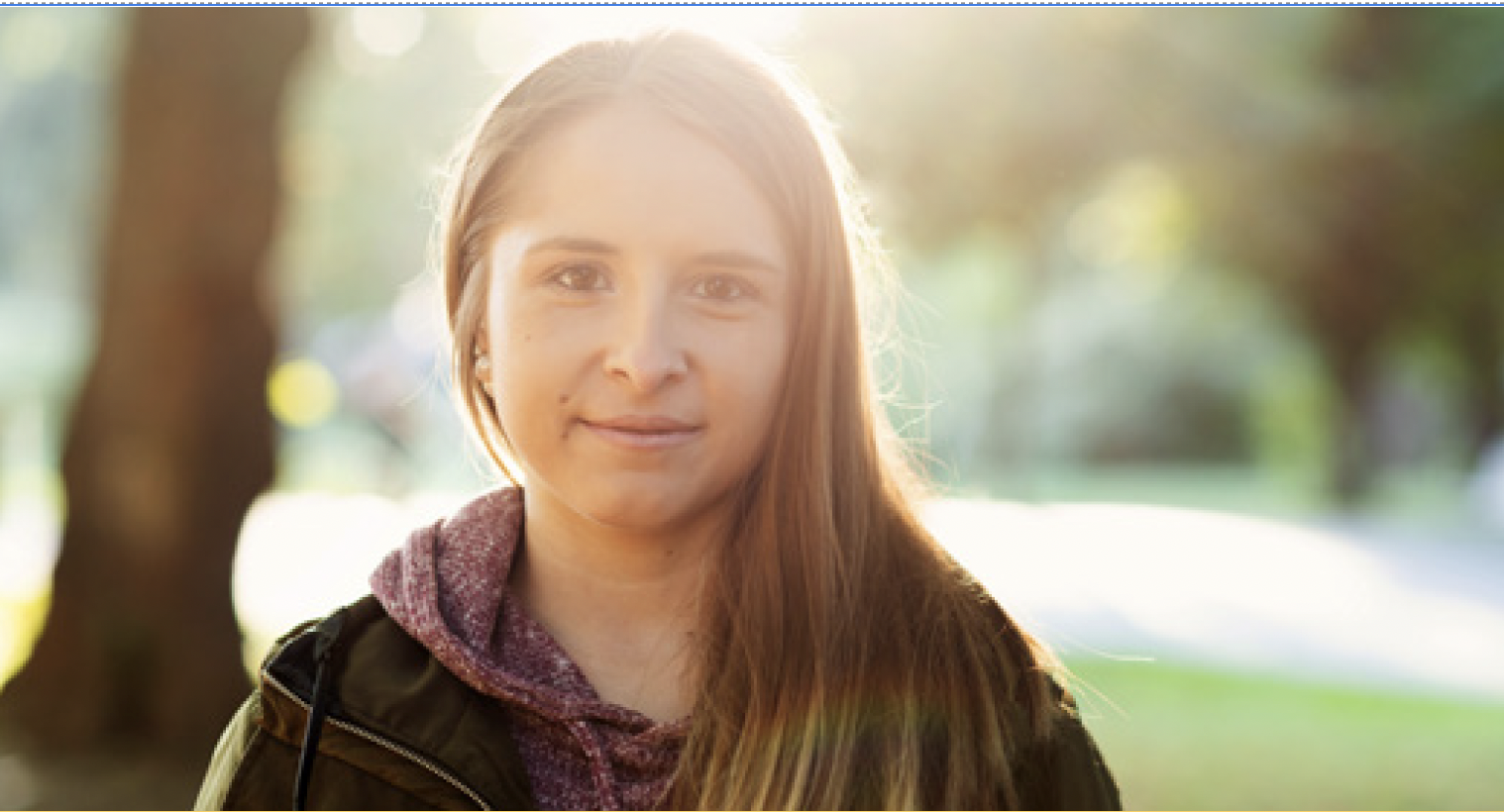 A young woman with long brown hair stands outdoors, smiling softly at the camera. She wears a hoodie and jacket. Sunlight shines behind her, creating a warm background—perfect for events for Virginia boys and girls among blurred trees and greenery.