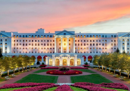 A large, elegant white hotel with tall columns and glowing lights sits behind landscaped gardens and flower beds at sunset, hosting special events for Virginia boys and girls beneath a pink and orange sky.