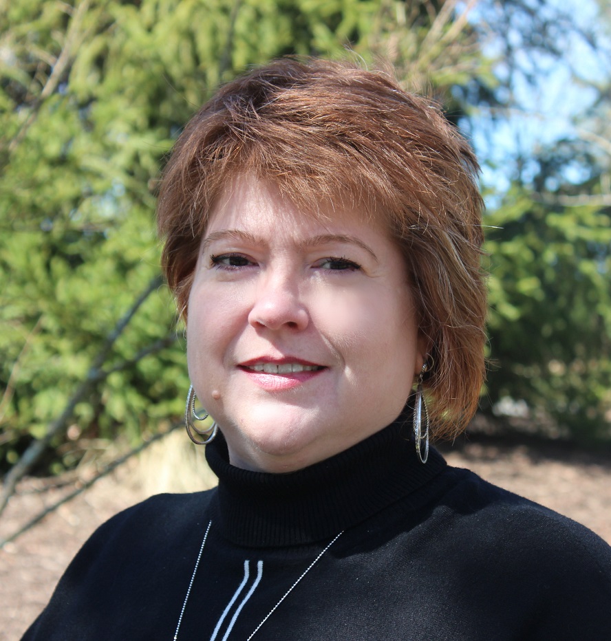 A woman with short auburn hair, wearing a black turtleneck sweater and silver earrings, stands outdoors in front of green trees, smiling slightly at the camera—representing Virginia education group homes services for boys and girls.
