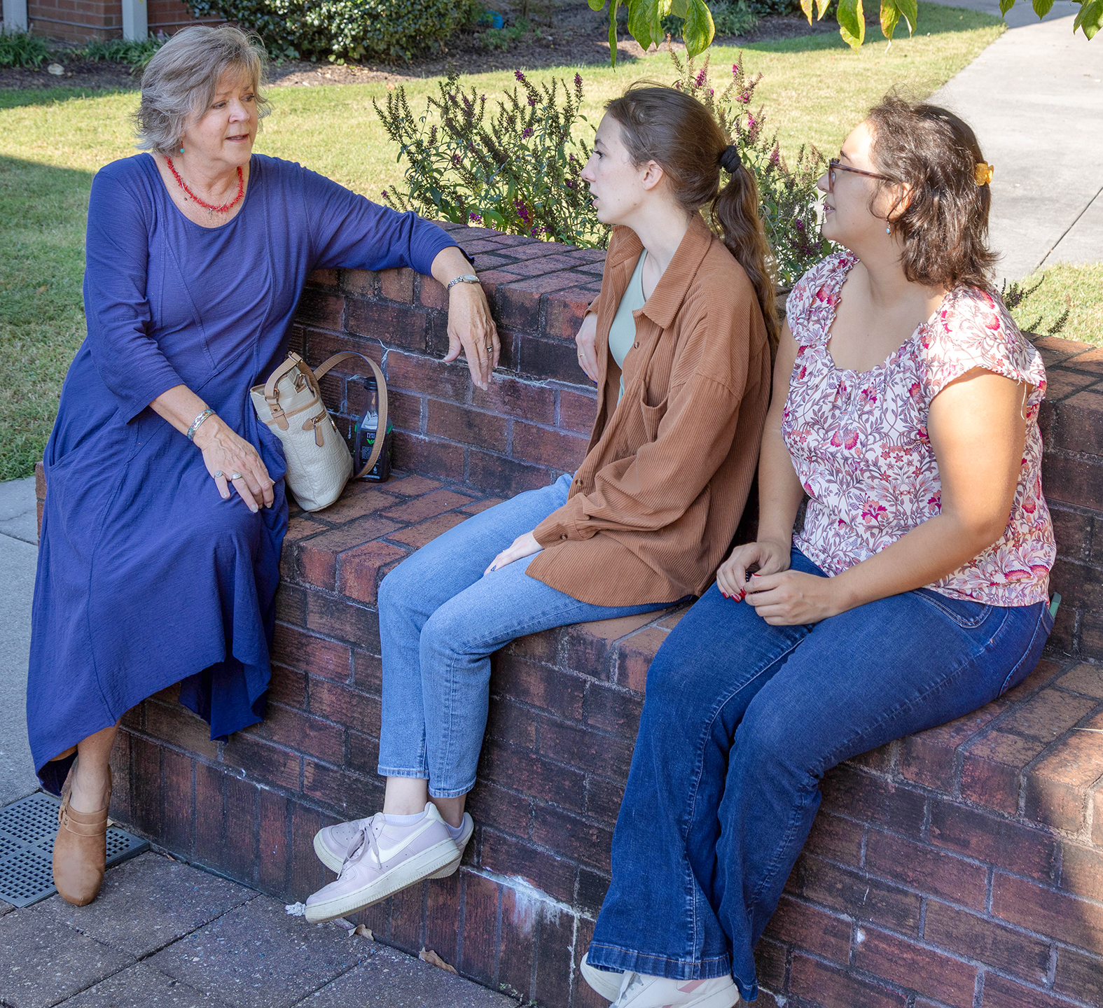 Three women sit on a brick wall outdoors, chatting about upcoming events for Virginia boys and girls. The older woman in a blue dress faces two younger women—one in a brown shirt and jeans, the other in a floral top—amid greenery and a sidewalk backdrop.
