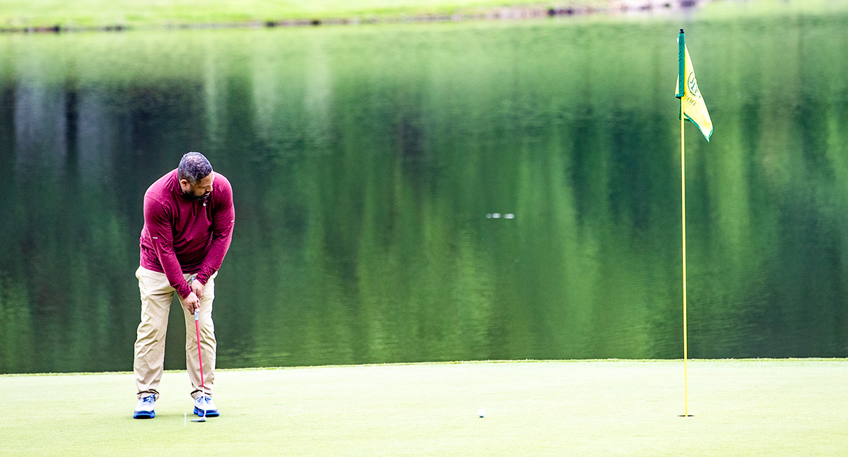 A man in a red sweater putts a golf ball on a green near a yellow flag, with a calm lake and green trees in the background—perfect scenery for events for Virginia boys and girls who love golf.