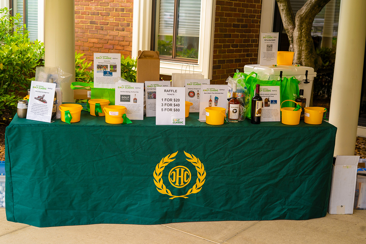 A raffle table covered with a green cloth displaying the JHC logo, featuring raffle prizes like bottles, buckets, and boxes. Signs list raffle prices: 1 for $20, 3 for $40, and 5 for $80—all supporting events for Virginia boys and girls.