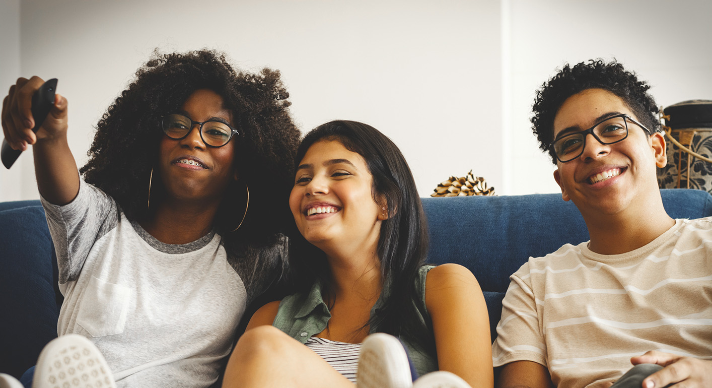Three smiling teenagers sit close together on a couch, laughing as they watch TV. One points a remote excitedly. They look like they're having fun, much like friends at events for Virginia boys and girls.