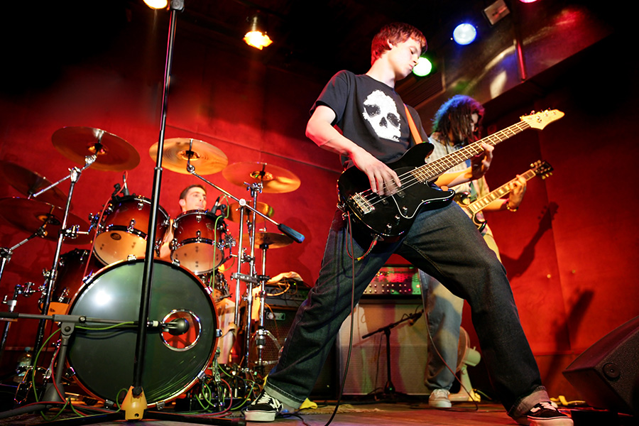A rock band performs on stage; a bassist in the foreground plays energetically, a drummer is behind a drum kit, and a guitarist stands to the right—perfect for exciting events for Virginia boys and girls. The lighting is vibrant with red and orange tones.
