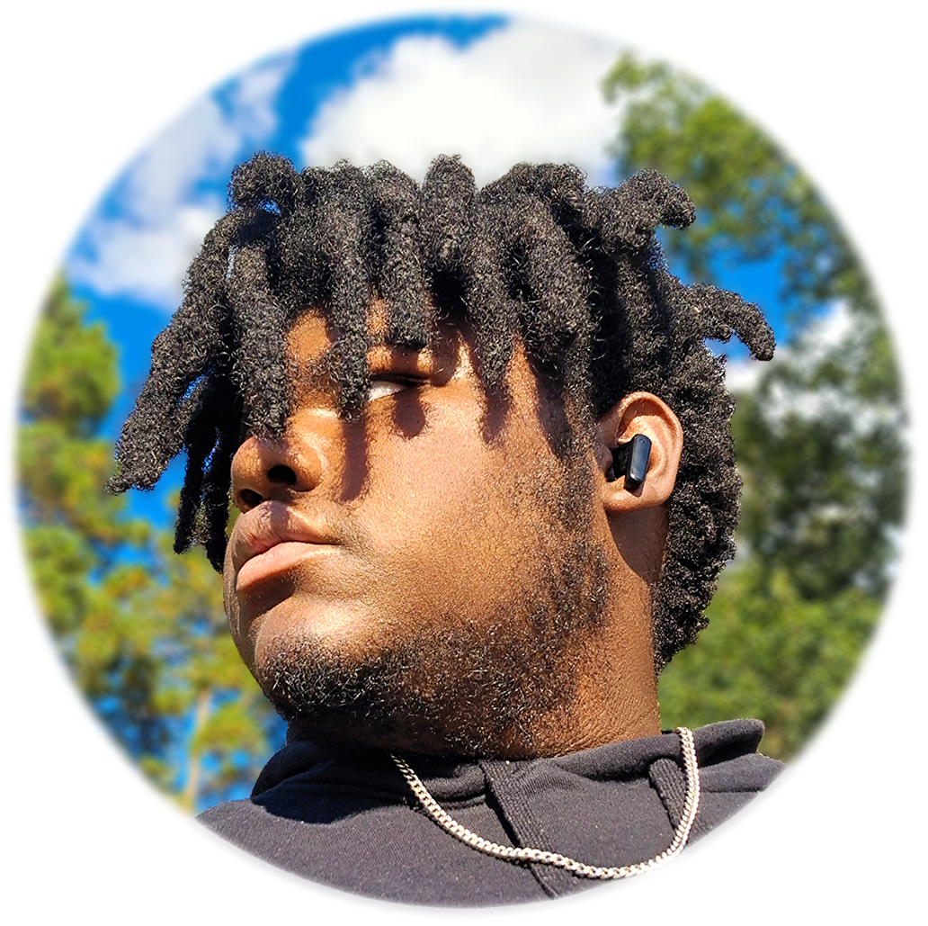 A person with short, thick twists in their hair, wearing a black hoodie, silver chain, and wireless earbud, looks to the side outdoors with trees and blue sky—a look of calm reflecting Virginia support for youth facing emotional and behavioral issues.