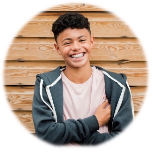 A smiling teenager with short curly hair and braces wears a pink shirt and a dark zip-up hoodie, standing in front of a wooden wall.