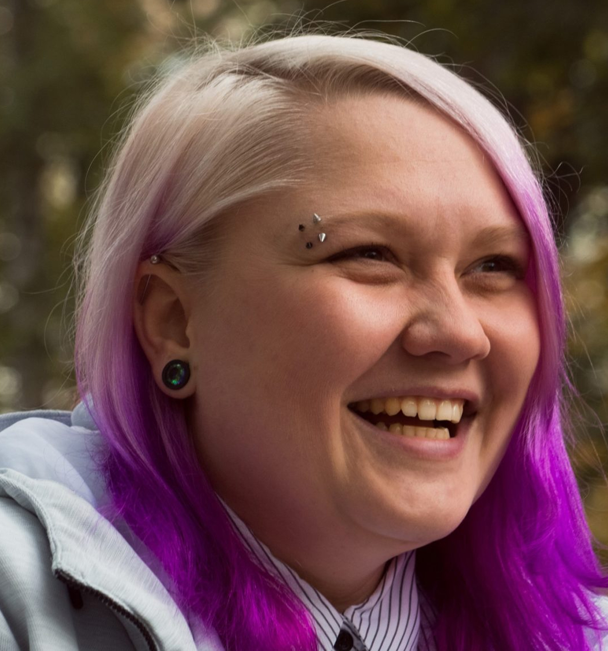 A person with light skin, light purple and pink hair, eyebrow piercings, and dark earrings is smiling broadly outdoors, wearing a light-colored jacket and striped shirt. Trees are blurred in the background.